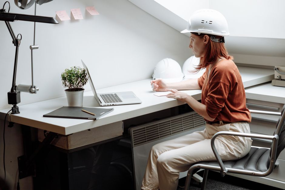 Caucasian woman in helmet working on a laptop in a minimalist office setting with a focus on architecture and design.